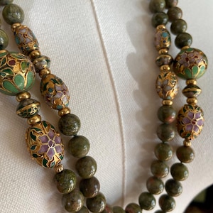 Puede incluir: Un collar de doble cadena con cuentas de piedra verde y marr&oacute;n y detalles florales de oro y esmalte.