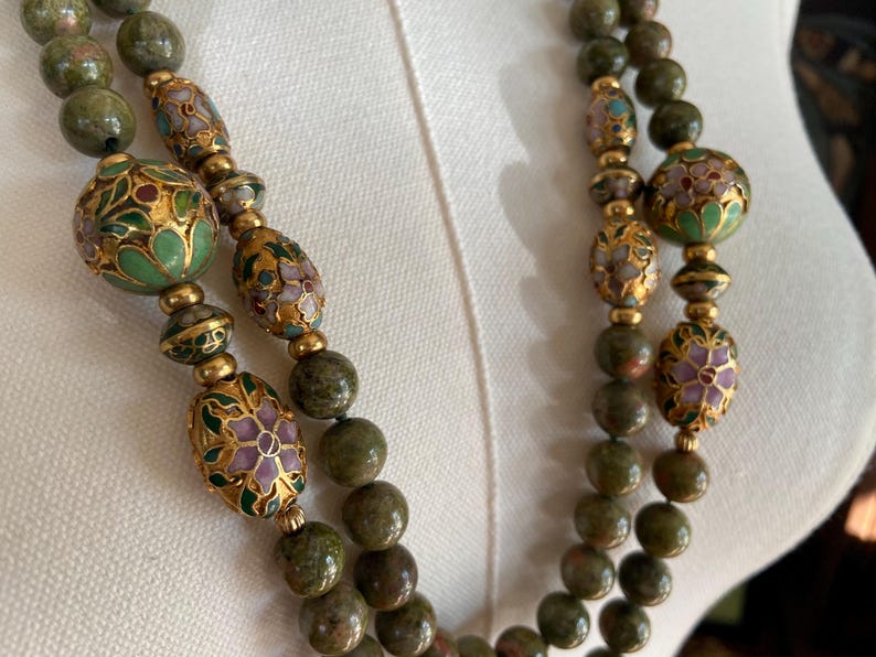 Puede incluir: Un collar de doble cadena con cuentas de piedra verde y marr&oacute;n y detalles florales de oro y esmalte.