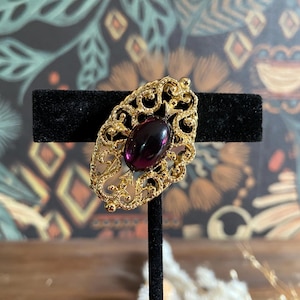 May include: A vintage gold-toned brooch with an oval, deep purple stone at its center. The brooch has an ornate, openwork design. The jewelry is displayed on a black stand against a patterned background.