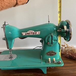 May include: A teal blue vintage sewing machine with a metal foot pedal and a silver needle. The machine has a white label that reads "*** UNITED" and another label that reads "1101 DE LUXE".