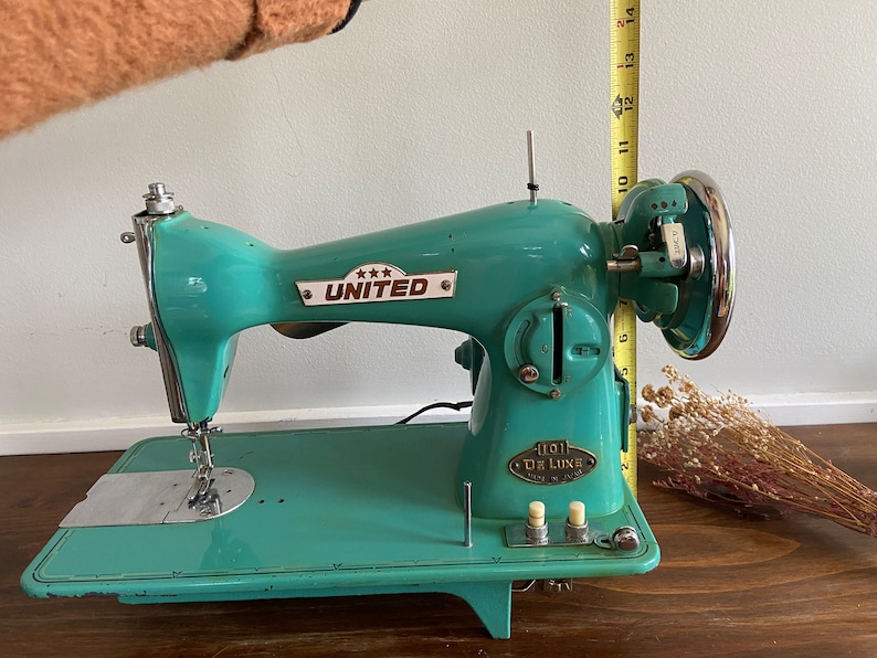 May include: A teal blue vintage sewing machine with a metal foot pedal and a silver needle. The machine has a white label that reads "*** UNITED" and another label that reads "1101 DE LUXE".