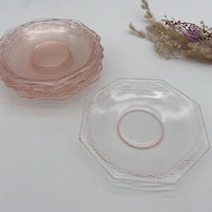 May include: A stack of pink glass saucers with a textured design and a single saucer with an octagonal shape. Dried flowers are in the background. The saucers are a light pink color.