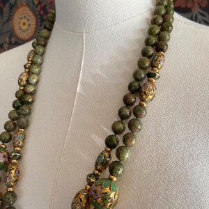 Puede incluir: Un collar de doble cadena hecho de cuentas de piedra verde y marr&oacute;n con detalles dorados y cuentas redondas m&aacute;s grandes, multicolores, con dise&ntilde;os florales.