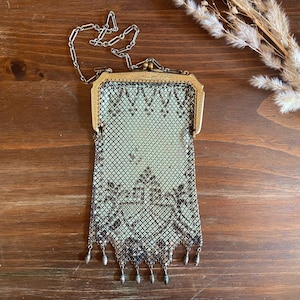 May include: A vintage metal mesh purse with a decorative frame and chain handle. The purse has a delicate, patterned design in black and silver, with small dangling accents at the bottom. The frame is gold-toned, adding a vintage aesthetic.