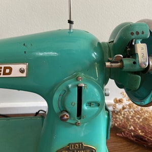 May include: A teal blue vintage sewing machine with a chrome foot pedal and a metal dial with the letters 'R', 'O', 'F' and '0-1'. The machine has a metal plate with the word 'UNITED' and three stars.