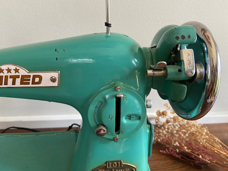 May include: A teal blue vintage sewing machine with a chrome foot pedal and a metal dial with the letters 'R', 'O', 'F' and '0-1'. The machine has a metal plate with the word 'UNITED' and three stars.