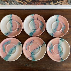 May include: Six ceramic saucers with a pink and white color scheme, accented with teal and navy blue brushstrokes. The saucers have a circular design with a central indentation for a cup. The set is arranged on a dark wooden surface.