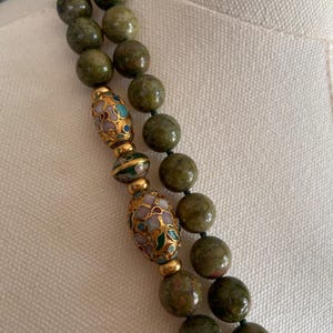 Puede incluir: Un collar de doble cadena hecho de cuentas de piedra verde con detalles dorados y de esmalte. Las cuentas son de forma ovalada y tienen un color verde moteado. Los detalles dorados son en forma de peque&ntilde;as cuentas y los detalles de esmalte son en forma de dise&ntilde;os florales.