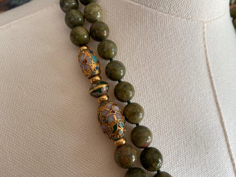 Puede incluir: Un collar de doble cadena hecho de cuentas de piedra verde con detalles dorados y de esmalte. Las cuentas son de forma ovalada y tienen un color verde moteado. Los detalles dorados son en forma de peque&ntilde;as cuentas y los detalles de esmalte son en forma de dise&ntilde;os florales.
