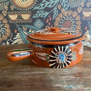 Puede incluir: Una pequeña olla redonda de cerámica de color terracota con tapa y asa. La olla está decorada con diseños florales pintados a mano en blanco, negro y azul. El asa tiene detalles verdes y marrones. La olla se utiliza probablemente para cocinar o servir.