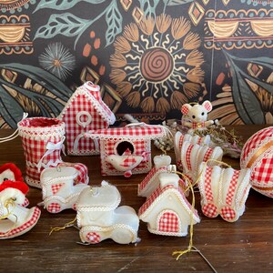 May include: A selection of Christmas decorations in a red and white gingham design. The ornaments include miniature houses, boots, a mouse figure, and a large bauble. The decorations are trimmed with white lace and ribbon. The background features a dark floral pattern.