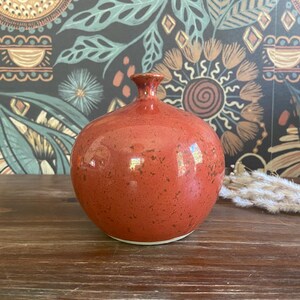 May include: A round, orange ceramic vase with a speckled texture. The vase has a narrow neck and a wide, rounded body. It sits on a wooden surface, with a patterned wallpaper background. Decorative dried plants are to the right of the vase.