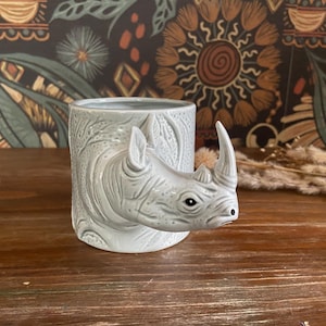 Może przedstawiać: Jasnoszary ceramiczny kubek z szczegółową głową nosorożca wystającą z boku. Kubek ma cylindryczny kształt z teksturowanymi wzorami liści. Nosorożec ma charakterystyczny róg i realistyczne szczegóły twarzy.