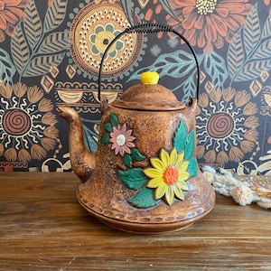 Puede incluir: Tetera de cerámica marrón con asa negra y pomo amarillo en la tapa. La tetera está decorada con diseños florales en relieve de colores, incluyendo un gran girasol amarillo y una flor rosa.