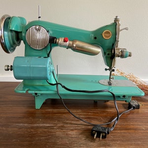May include: A vintage turquoise blue sewing machine with a metal foot pedal and a black electrical cord. The machine has a silver bobbin winder and a silver needle.