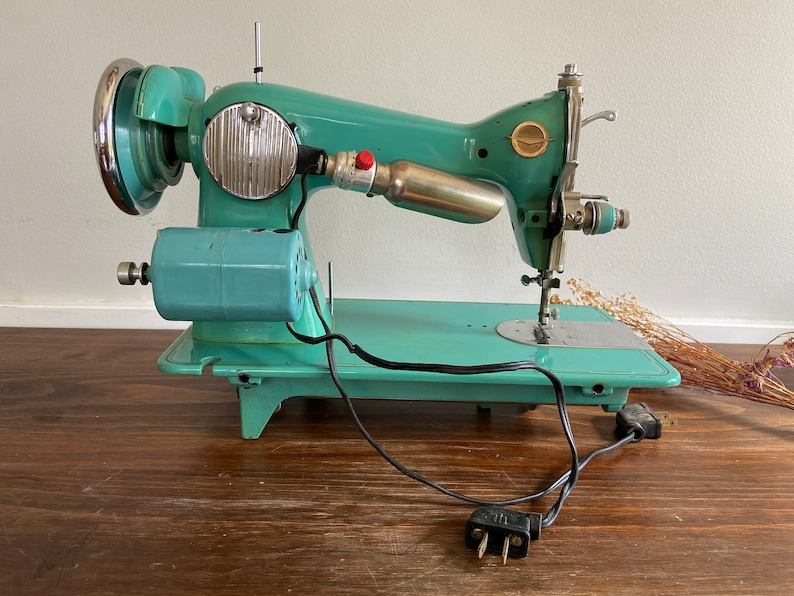 May include: A vintage turquoise blue sewing machine with a metal foot pedal and a black electrical cord. The machine has a silver bobbin winder and a silver needle.