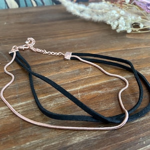 May include: A layered choker necklace with a black suede band and a delicate rose gold chain. The necklace is displayed on a wooden surface, showcasing its elegant design. The jewelry is a stylish accessory.