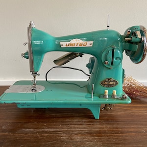 May include: A vintage turquoise blue sewing machine with the brand name "UNITED" and "1101 DE LUXE" printed on the side. The machine has a silver needle and foot pedal.