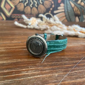Puede incluir: Un reloj de pulsera vintage con esfera oscura y bisel plateado. El reloj tiene una correa de cuero verde turquesa. El reloj está sobre una superficie de madera. Un accesorio único.