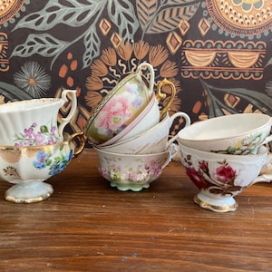 May include: A collection of vintage teacups with floral designs. The cups feature gold trim and delicate patterns in shades of pink, blue, and purple. Some cups are stacked, showcasing their varied shapes and sizes.