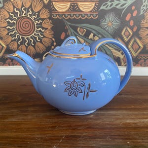 May include: A light blue teapot with a gold lid and gold floral designs. The teapot is sitting on a wooden surface.
