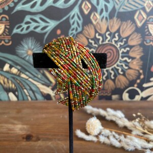 May include: A multi-strand bracelet featuring small, colorful beads in shades of red, yellow, green, and gold. The bracelet is displayed on a black stand against a backdrop of a floral patterned wallpaper.