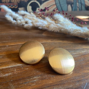 May include: Two gold-colored, round earrings with a textured, brushed metal finish. The earrings are set on a wooden surface, with dried decorative plants in the background. The earrings have a simple, classic design.