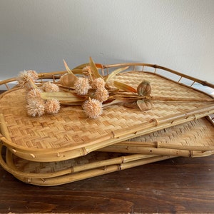 May include: Two woven bamboo trays with rounded edges, stacked on top of each other. The top tray has a dried flower arrangement with small, round, beige flower heads.