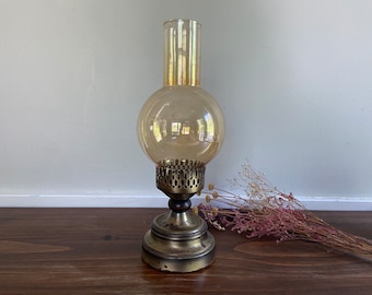 May include: A vintage brass candle holder with a clear glass globe. The holder has a decorative design and is sitting on a wooden surface.