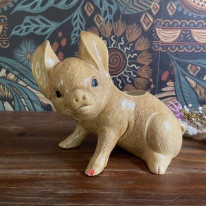 May include: A tan ceramic pig figurine with large ears and a small snout. The pig is in a crouching position, with its front legs extended. The figurine has black eyes and a small red mark on one foot. The background is a dark floral pattern.