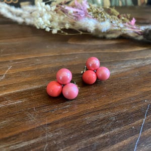 Puede incluir: Un par de pendientes vintage de clip. Cada pendiente presenta tres cuentas redondas de color coral y rosa. Los pendientes están colocados sobre una superficie de madera oscura, con flores secas en el fondo. Un accesorio clásico.