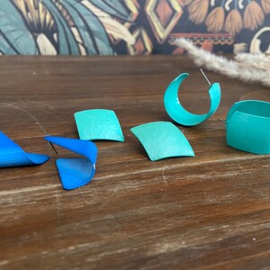 May include: A collection of colorful earrings in shades of blue and teal. The earrings include hoop, square, and curved designs. The earrings are displayed on a wooden surface.