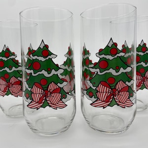 May include: A set of four clear drinking glasses. Each glass is decorated with a green Christmas tree design, red baubles, and a red and white striped bow. The glasses are tall and cylindrical.