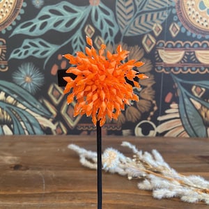 Puede incluir: Un objeto decorativo naranja brillante, que se asemeja a una flor, montado sobre un soporte negro. El objeto está compuesto por numerosos elementos pequeños y translúcidos de color naranja. El fondo presenta un papel pintado estampado en tonos turquesa, marrón y crema. Se exhibe sobre una superficie de madera.