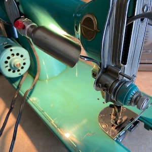 May include: A vintage turquoise sewing machine with a silver metal arm and a black cord. The machine has a light on the arm and a dial with numbers 1-12.