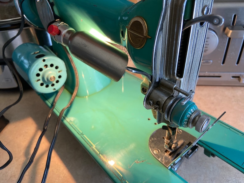 May include: A vintage turquoise sewing machine with a silver metal arm and a black cord. The machine has a light on the arm and a dial with numbers 1-12.