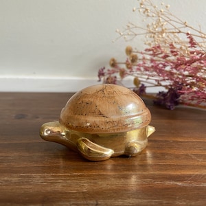 May include: A gold turtle trinket box with a brown and gold patterned lid. The box is sitting on a wooden surface.