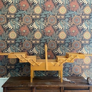 May include: A wooden sewing box with multiple compartments and a handle. The box is open, revealing storage spaces. It is set on a dark wooden table against a patterned floral wallpaper background.