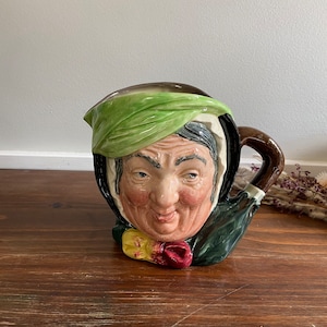 Puede incluir: Una jarra de cerámica con forma de cabeza de mujer con un pañuelo verde y un asa marrón. La mujer tiene una cara pintada con una sonrisa y un pañuelo verde.