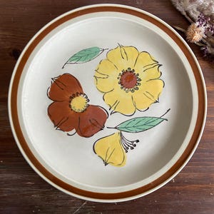 May include: A white ceramic dinner plate with a brown rim features a stylized floral design. The flowers are yellow, orange, and brown with green leaves.