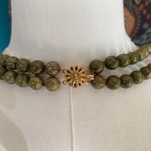 Puede incluir: Un collar de doble cadena hecho de cuentas verdes y marrones moteadas con un cierre dorado en forma de flor.