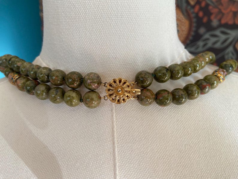 Puede incluir: Un collar de doble cadena hecho de cuentas verdes y marrones moteadas con un cierre dorado en forma de flor.