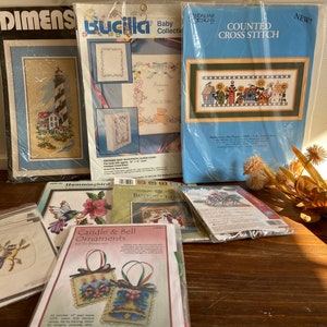 Puede incluir: Una colección de patrones de punto de cruz con varios diseños, incluyendo un faro, una cubierta de álbum para bebé, una fila de casas, un colibrí, un adorno navideño y un pájaro en una rama. Los patrones están empaquetados en plástico y etiquetados con las marcas "Dimensions", "Bucilla" y "Landmar Designs".