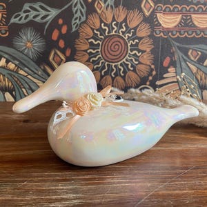 Puede incluir: Figura de pato de cerámica iridiscente con acabado nacarado. El pato presenta un arreglo decorativo de rosas y encaje color melocotón. Un artículo de decoración del hogar único.