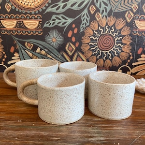 Könnte beinhalten: Vier gesprenkelte Keramikbecher in einer neutralen Farbpalette. Zwei Becher haben Henkel, zwei sind ohne Henkel. Die Becher sind auf einer Holzoberfläche mit einem dekorativen floralen Hintergrund angeordnet.
