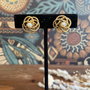 May include: A pair of gold-toned earrings in a vintage style. Each earring has a woven, floral design with a small, round, pearl-like centre. The earrings are displayed on a black stand against a patterned background.