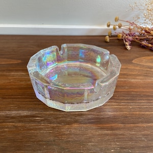 May include: Iridescent glass ashtray with a textured, faceted design. The ashtray has a round shape with four notches for resting cigarettes. The glass reflects a rainbow of colors. A decorative item for a home.
