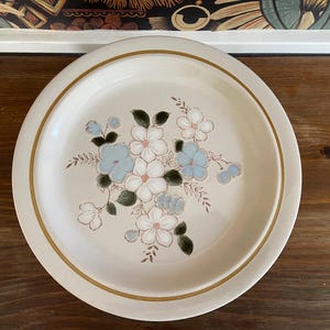 May include: A white ceramic dinner plate with a brown rim and a floral design featuring blue and white flowers. The flowers are arranged in a cluster and have green leaves.