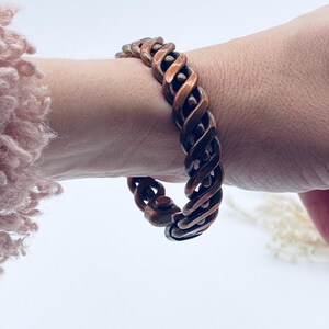 May include: A copper-toned bracelet with a twisted rope design. The bracelet is worn on a wrist, showcasing its intricate pattern and metallic sheen. The jewelry piece is a statement accessory.