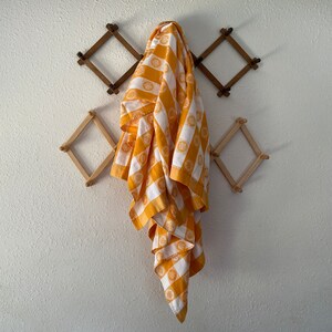May include: A white and orange patterned towel hangs on a wooden wall-mounted rack. The towel features a repeating pattern of orange circles and stripes. The rack is made of diamond-shaped wooden pieces.